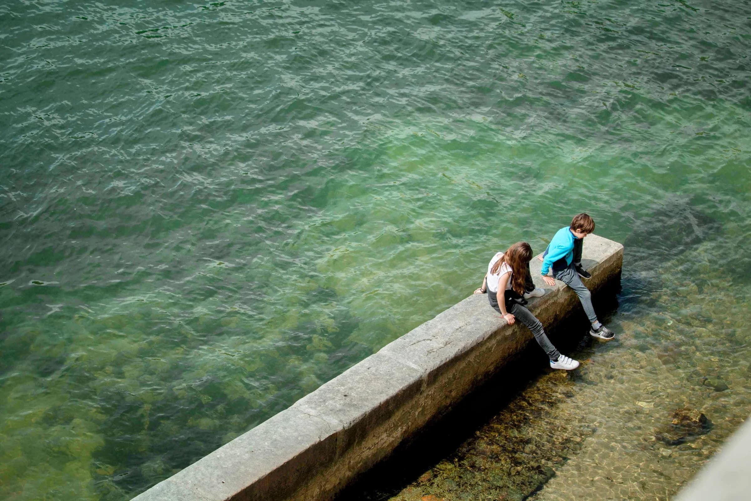 Two people sitting on a stone wall above clear green water.